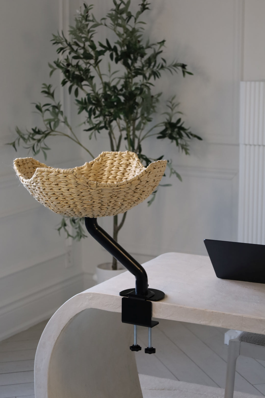 Empty handwoven cat bed from ERGO PURRCH™, mounted on a black stand attached to a white desk in a modern home office with a potted plant and a laptop in the background.