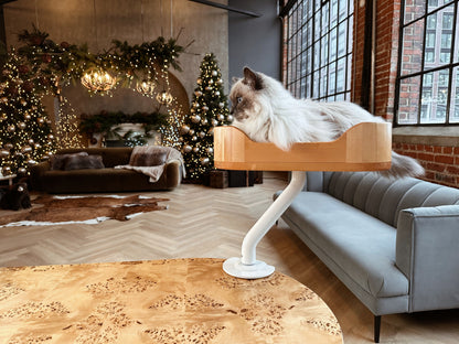 Beautiful Siberian cat resting in a wooden ERGO PURRCH™ desk-mounted cat bed with a white arm, in a stylish loft decorated with holiday lights, greenery, and a festive Christmas tree.